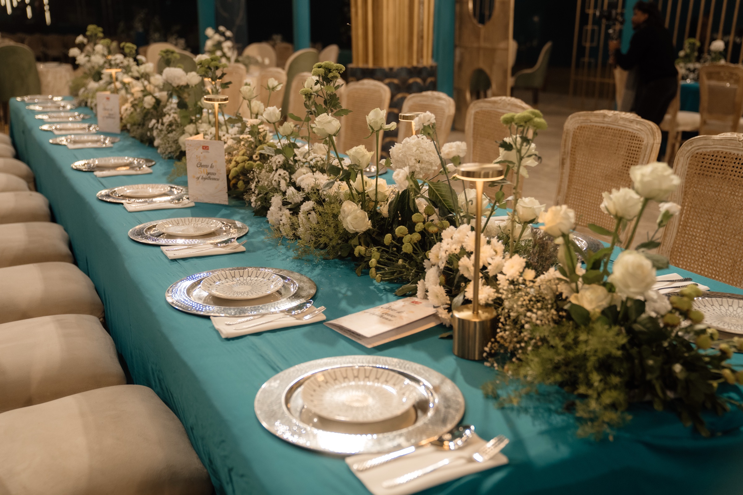 Long teal banquet table with white flowers and a card reading Cheers to 50 years of brilliance, Kabra golden anniversary